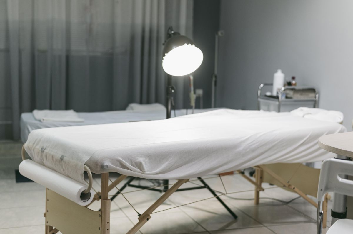 Kozmetički krevet prekriven belom podlogom u salonu lepote, sa lampom za tretmane i pomoćnim kolicima za pribor